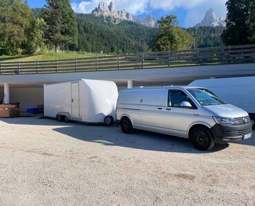 Ein grauer VW Bus mit einem weißen großen Anhänger steht auf einem tiefergelegenen Sandparkplatz. Darüber führt ein Weg oder Straße mit Holzgeländer.