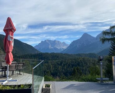 Eine Aussichtsplattform in den Bergen. Sicht auf grüne Wälder und Berge. Links eine Terrasse mit Tisch und Stühlen und geschlossenen roten Sonnenschirmen.