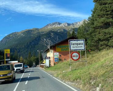 Fotografiert aus einem Auto mit Blick auf einer geraden Straße in den Bergen. Am rechten Straßenrand ein Schild mit der Aufschrift "Fernpass".